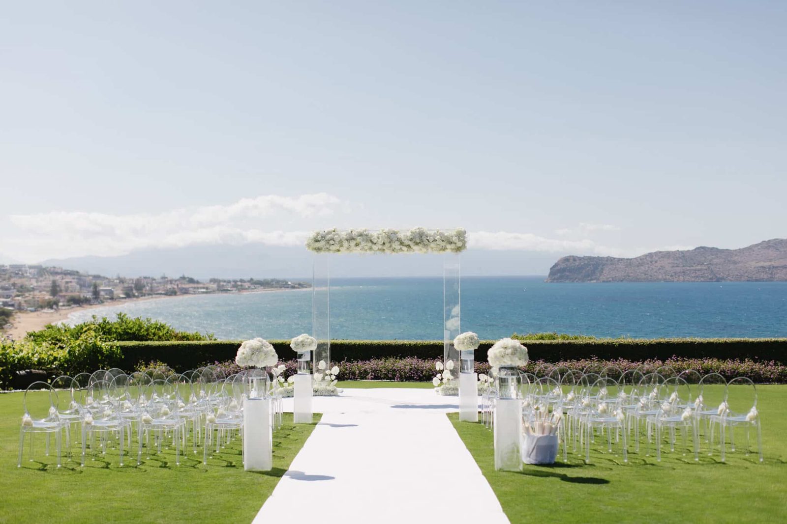 Clear ghost chairs and a white floral arch on a lawn above the sea in Greece